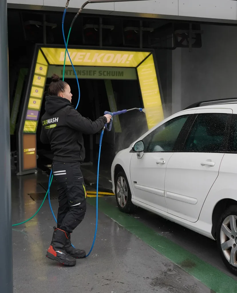 Een vrouw spuit een witte auto schoon bij een autowasstraat.